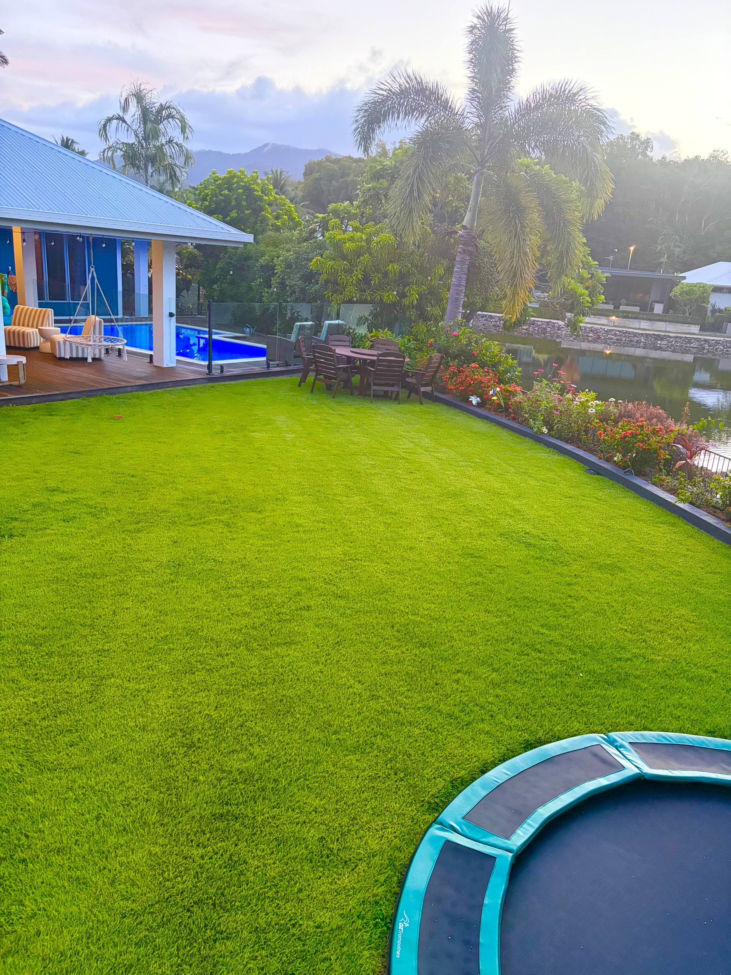 Fully fenced backyard lawn with in-ground trampoline, pool deck and lagoon at sunrise